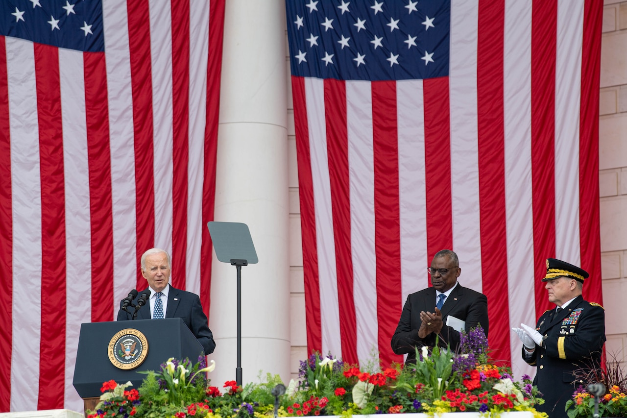 President Joe Biden speaks from a podium as the defense secretary and chairman applaud nearby with American flags hanging in the background.