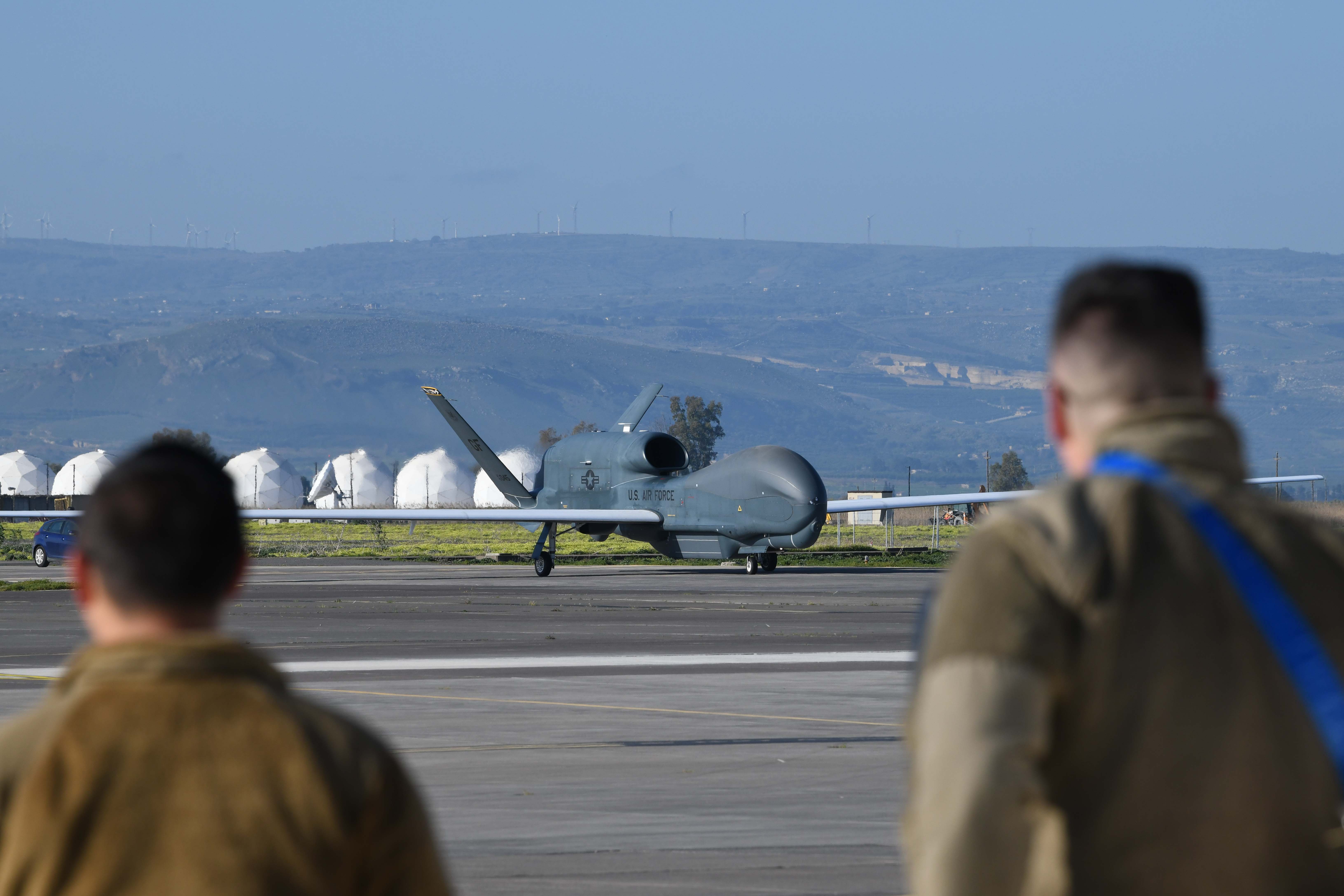 Two male airmen in brown jackets stand in front of a grey aircraft and blue sky.