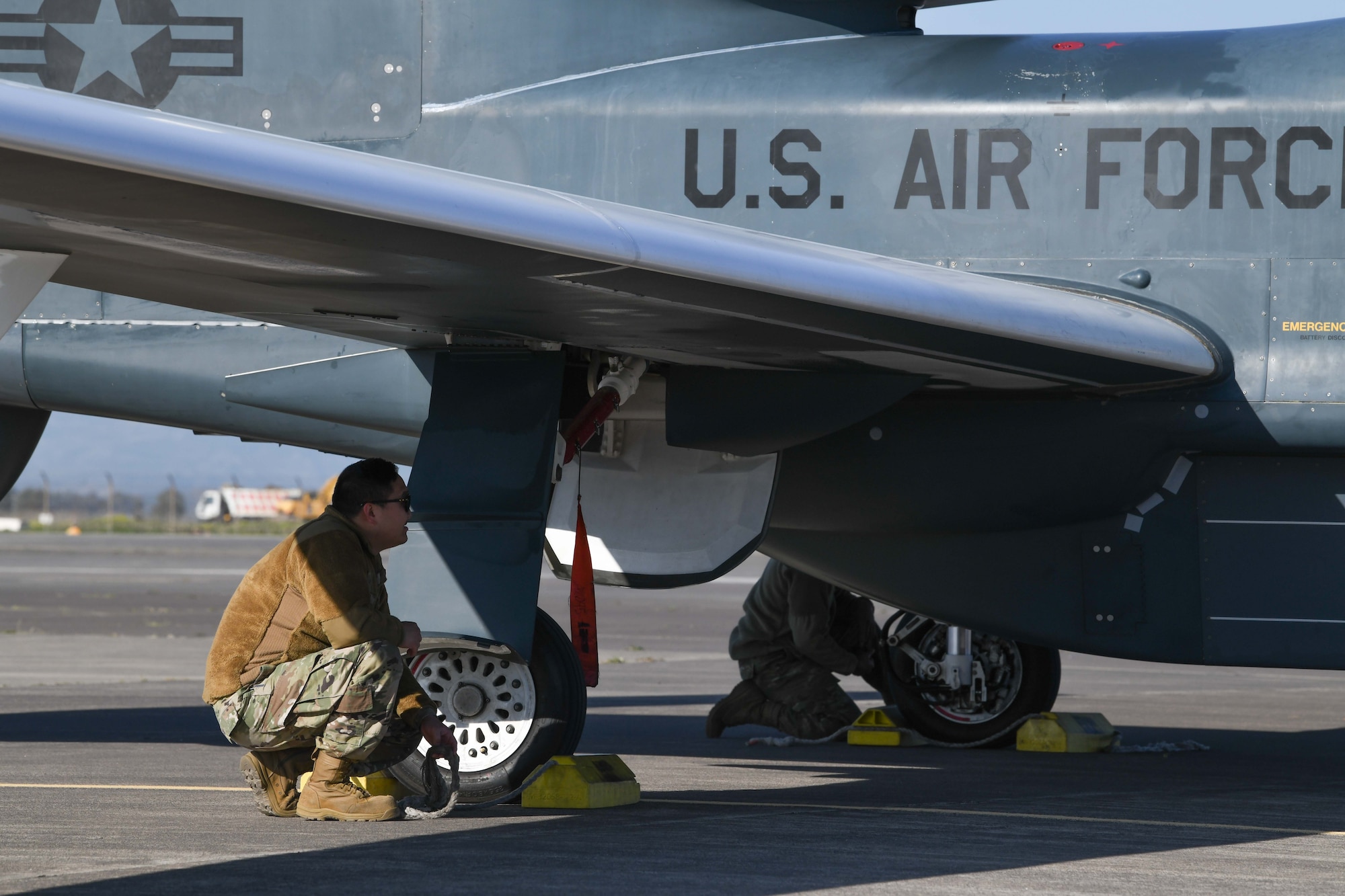 A male airman in a brown jacket and green pants kneels under the wing of a grey aircraft.