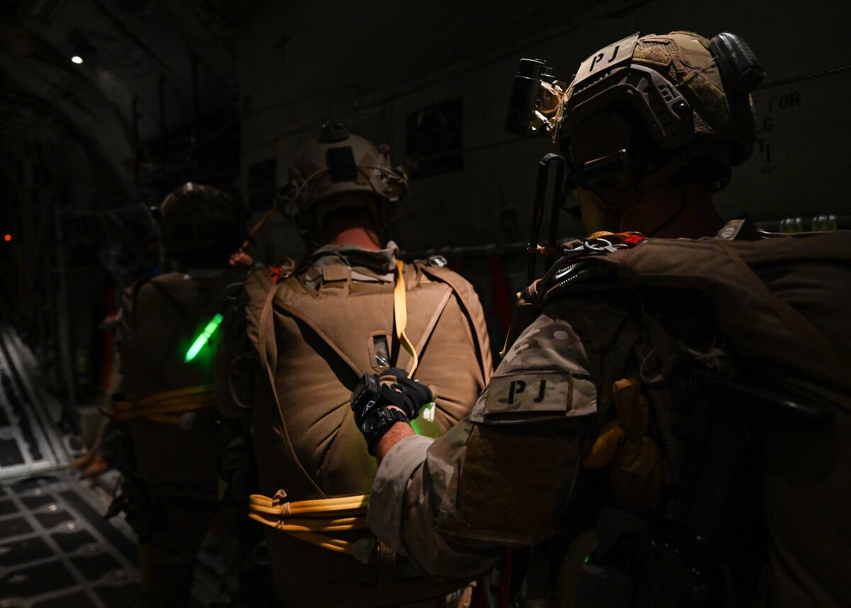 Men in military uniforms and gear check each other's gear in preparation for parachuting out of an airplane.