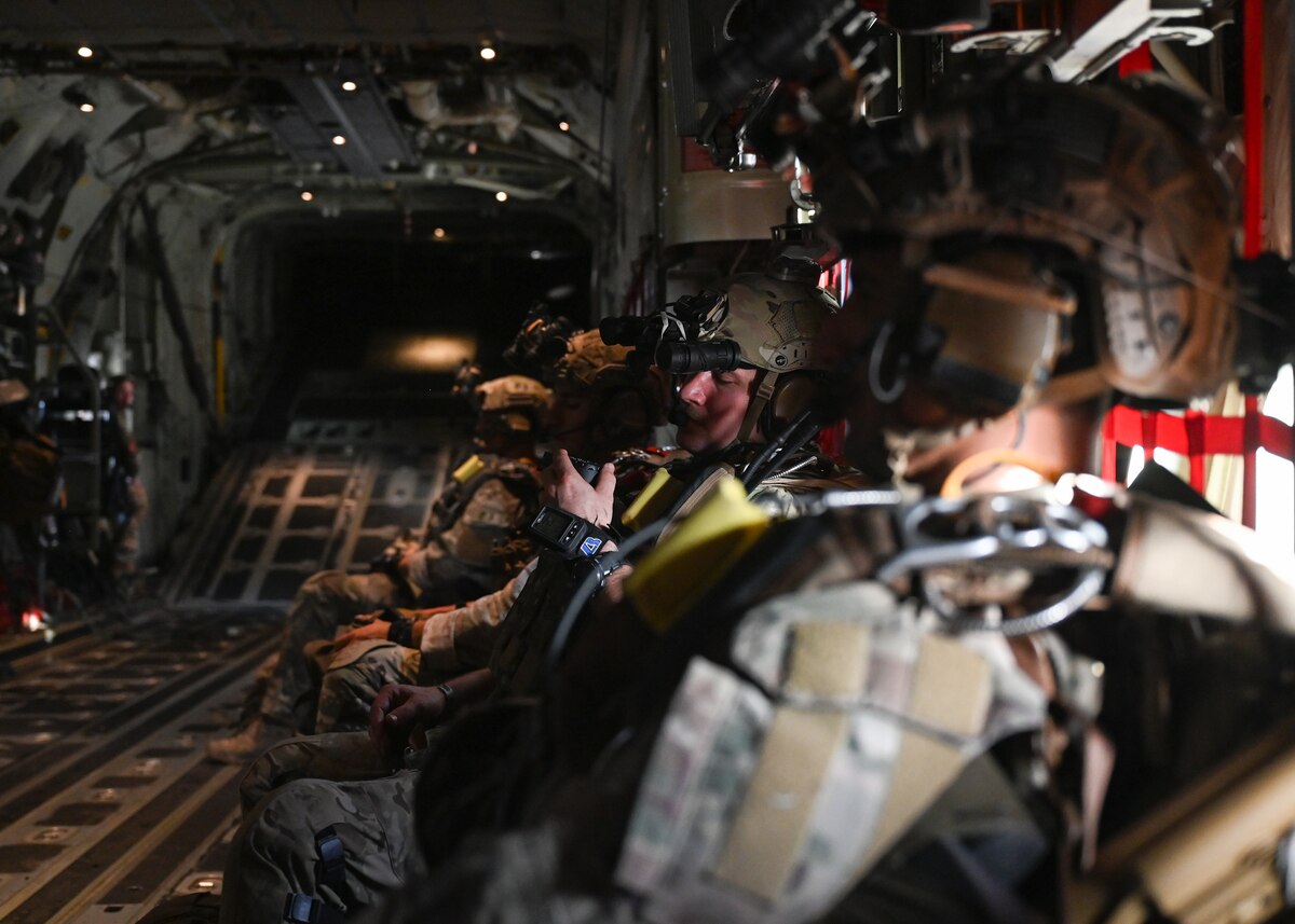Men in military uniforms and gear sit inside an aircraft in preparation for parachuting out of it.