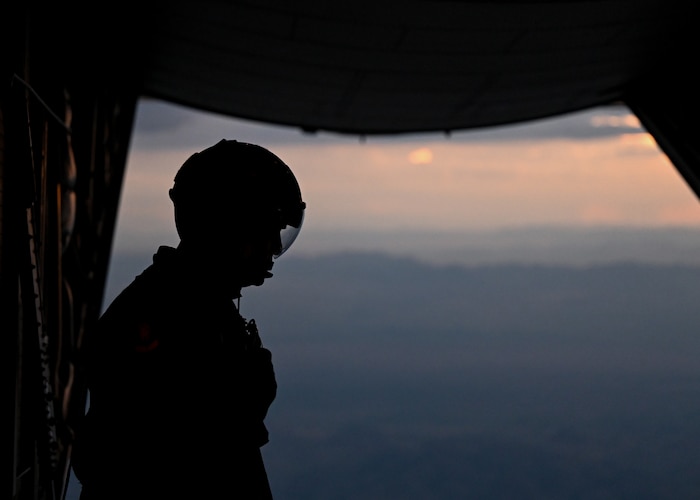 A man in a military uniform stands in the back of an aircraft with the back door open in preparation for pararescuemen to parachute out of.