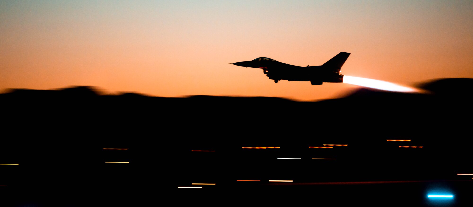 A silhouette of an F-16 Fighting Falcon soars over a silhouette of Arizona mountains with a sunset behind it, the F-16 has its after burner engaged.