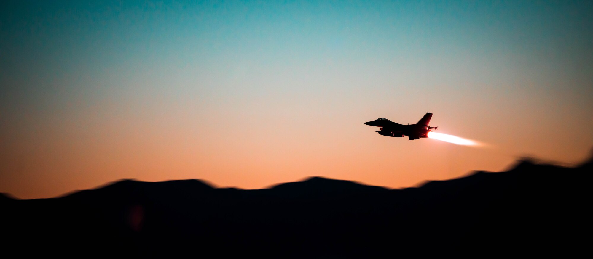 A silhouette of an F-16 Fighting Falcon soars over a silhouette of Arizona mountains with a sunset behind it, the F-16 has its after burner engaged.