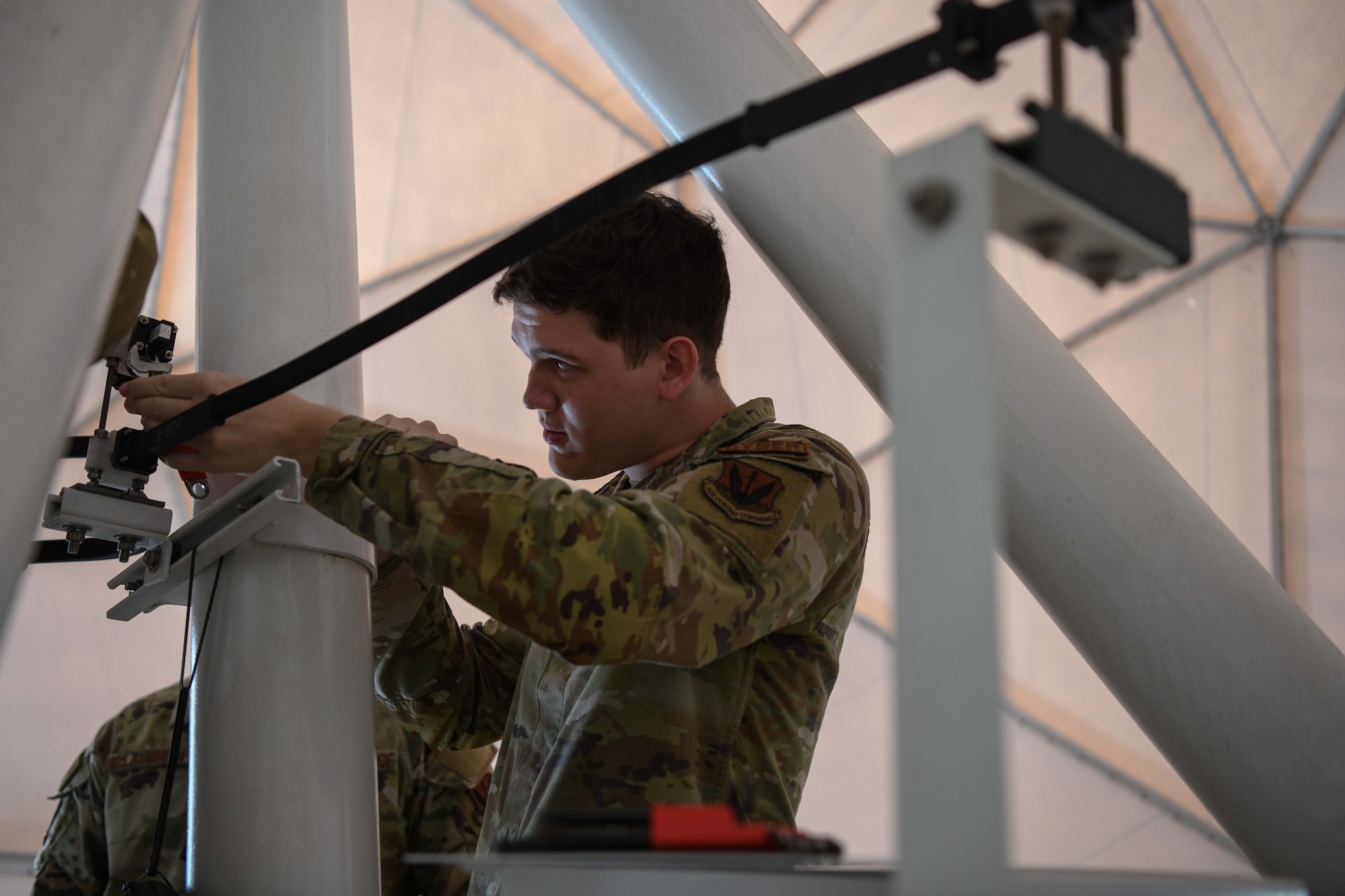A male airman in a green uniform conducts maintenance on a white beam.