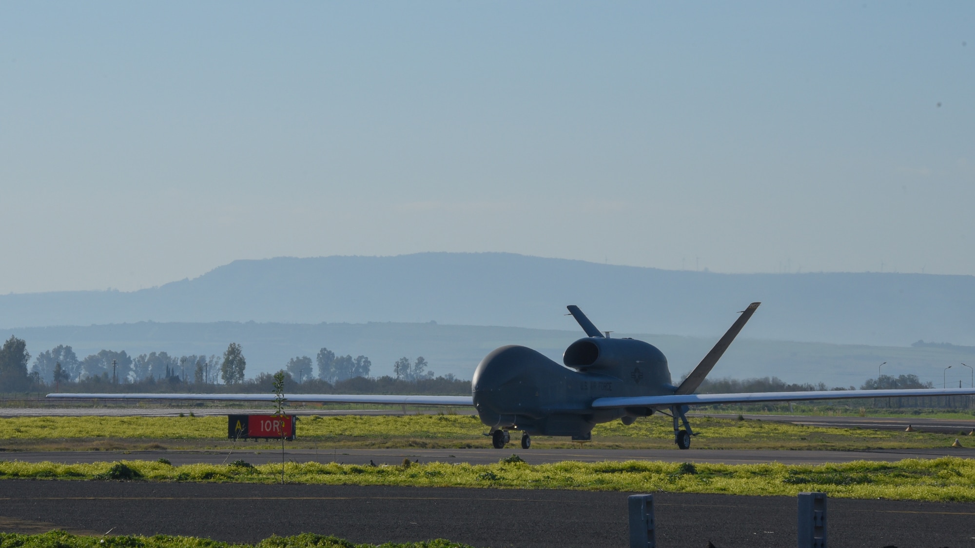 A grey aircraft is stopped in front of a blue sky and green grass is behind and in front of the tires.