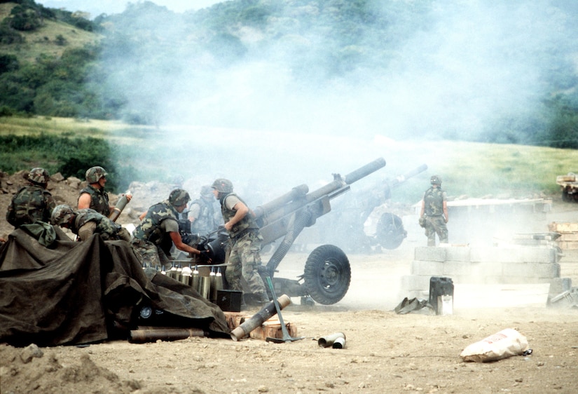 Soldiers fire a howitzer on dirt terrain with mountains in the background.