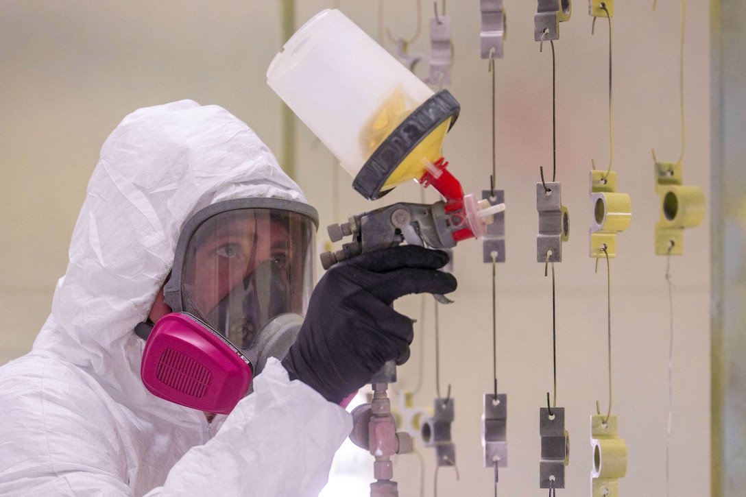 An airman in protective gear paints small airplane parts.