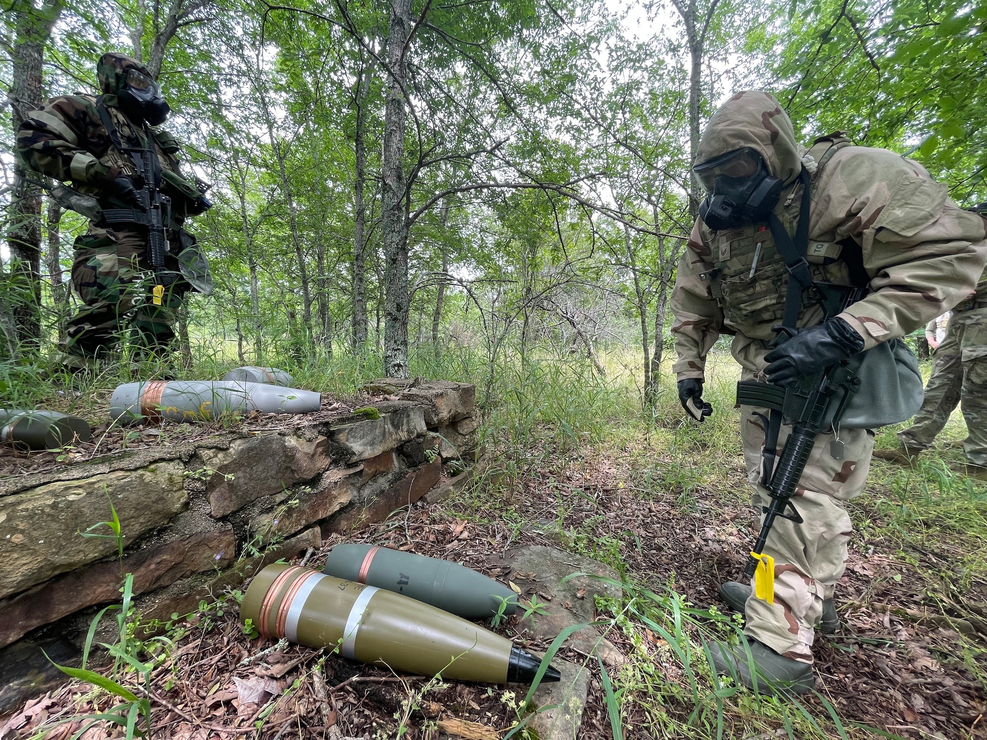 Two individuals in MOP4 gear stare down at a pile of explosives in the forest.