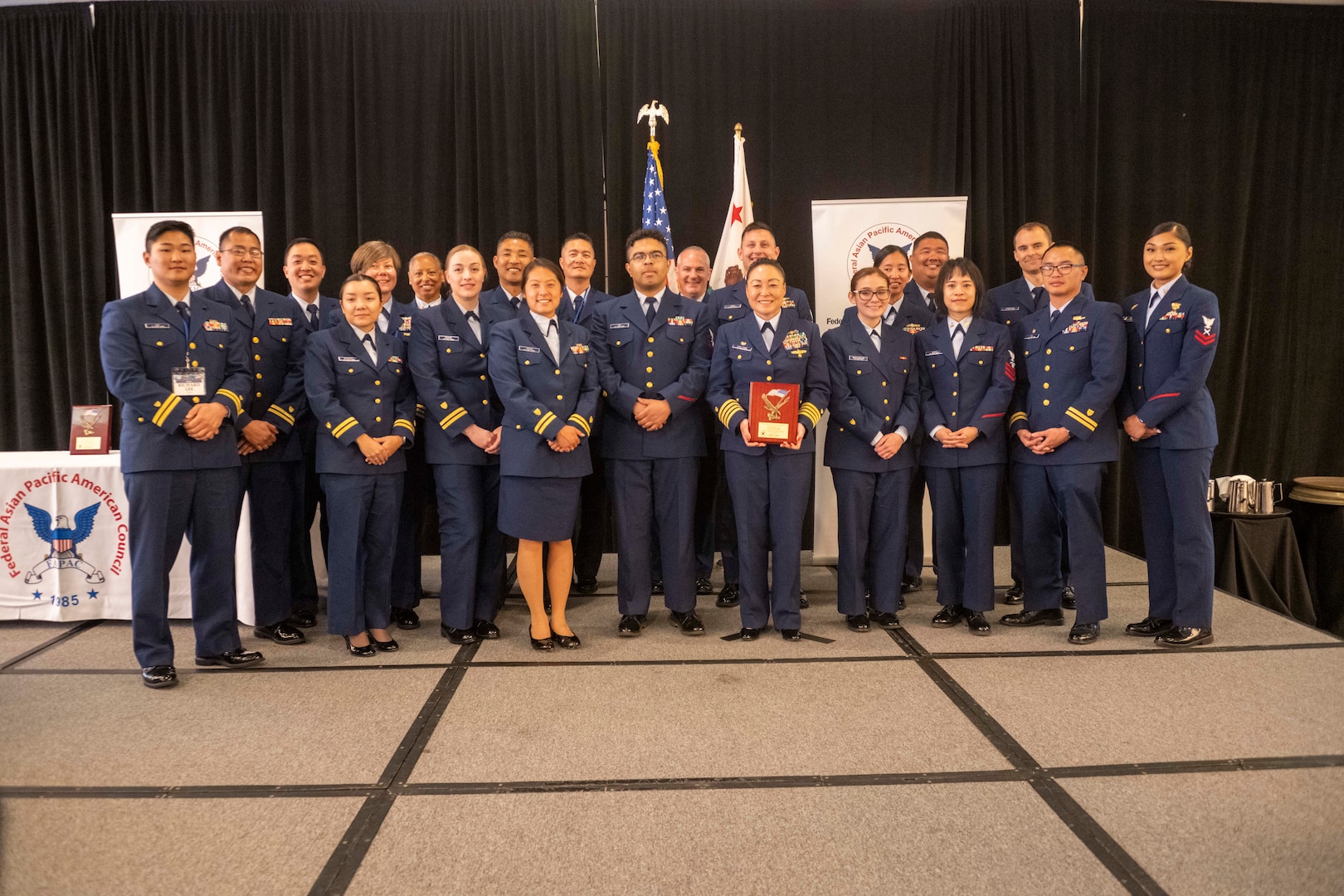 Members of the USCG Federail Asian Pacific American Council line up in two rows to pose for a picture. Capt. Eva Van Camp is in the front row holding her award.