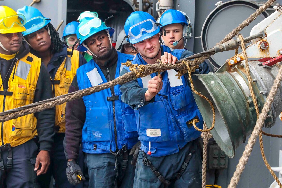 A sailor removes rope from a piece of equipment as other sailors watch.