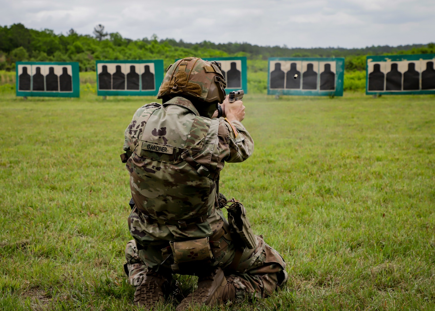 A Soldier kneels in a field facing multiple targets while shooting a pistol.