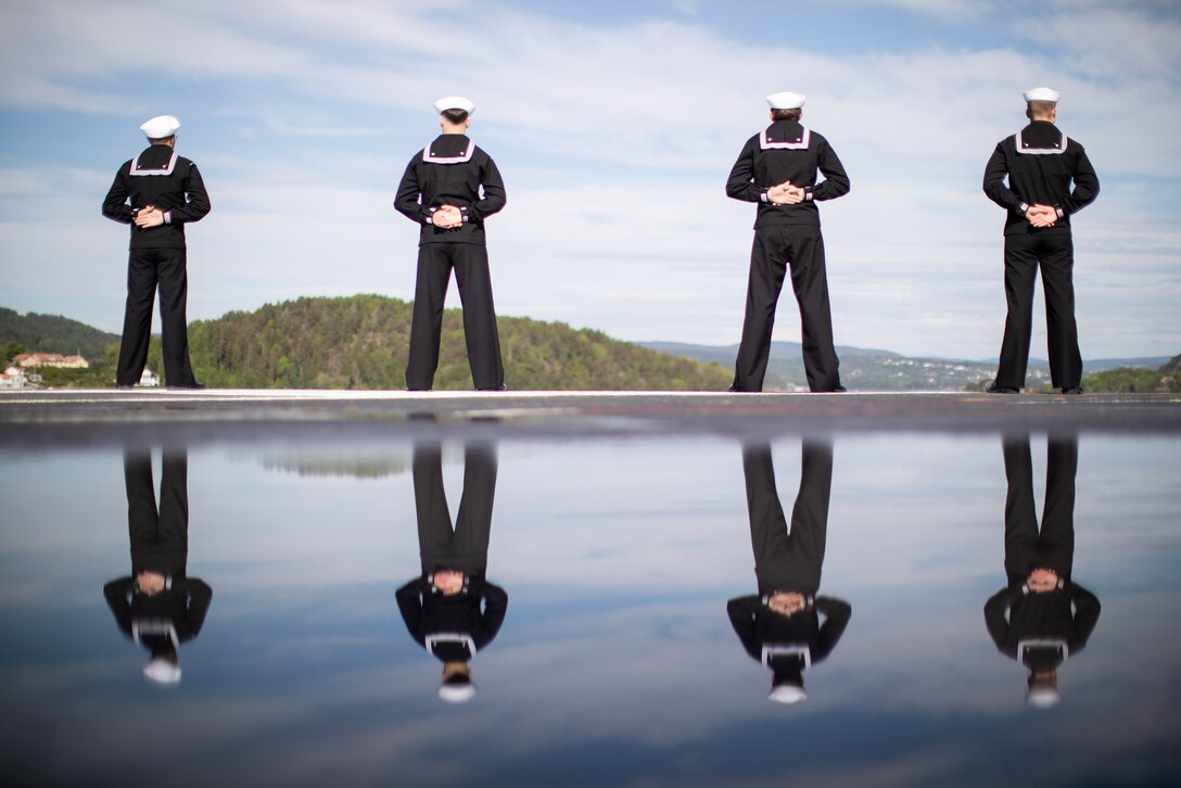 Four sailors, shown from behind and reflected in water, stand in a line, looking out onto a hilly area.