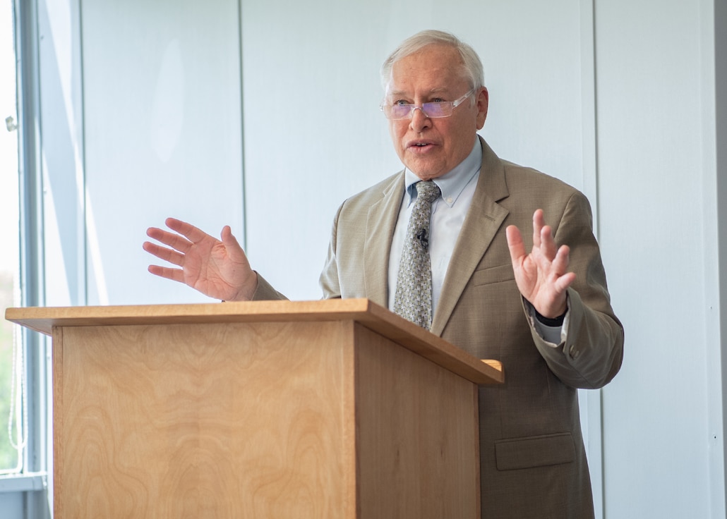 A man in a tan suit speaks from behind a podium