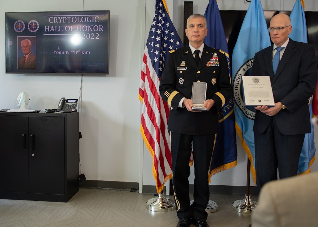 A mounted TV displays the name Youn P. Kim. Two men, one in military uniform and one in a suit, stand solemnly.