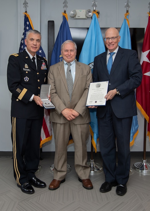 From left to right stand a man in military uniform, a man in a tan suit, and a man in a gray suit.