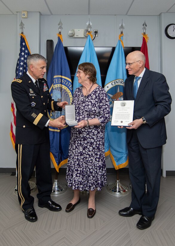 From left to right stand a man in military uniform, a woman in a purple floral dress, and a man in a suit. The first man hands the woman a plaque.