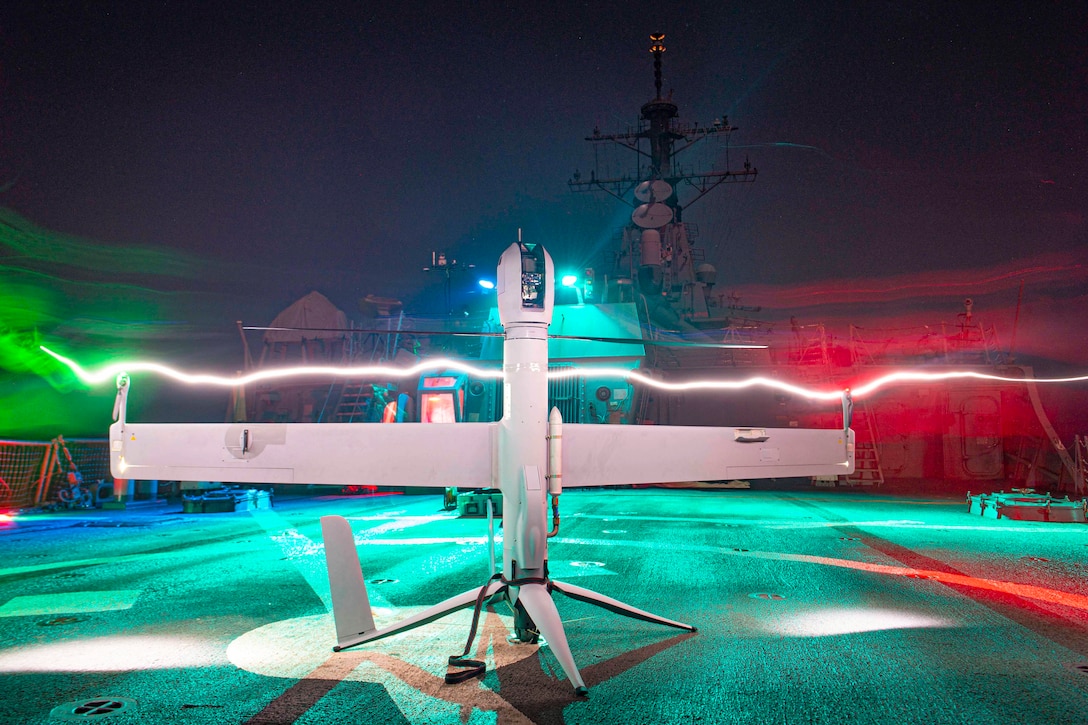 An unmanned aerial vehicle sits on the flight deck of a ship as a streak of light passes near the center of it illuminated by red, blue, green and white lights.
