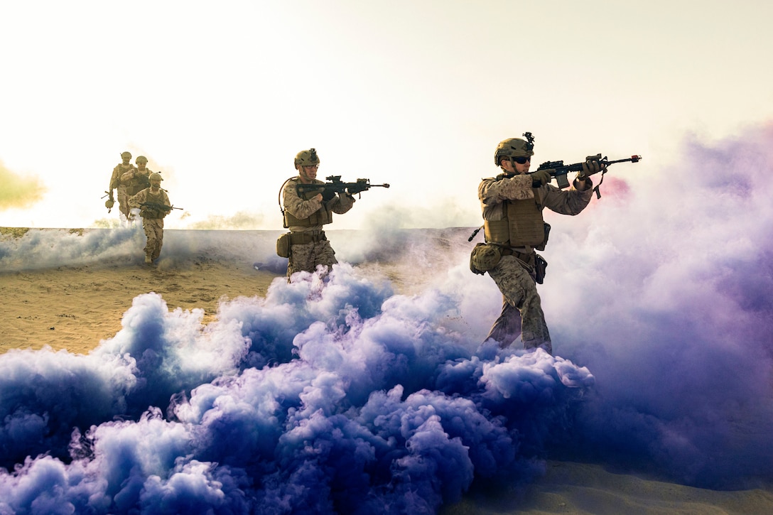 Marines aiming weapons walk in a line through clouds of purple smoke in a field.