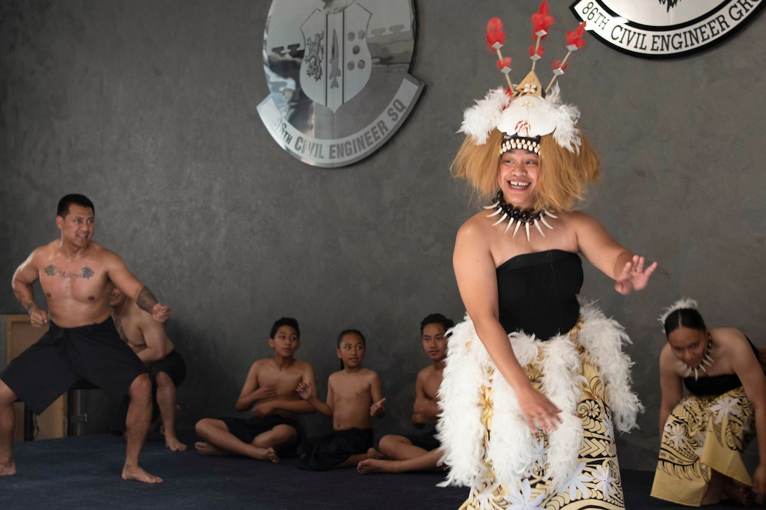 Airmen wearing traditional Samoan clothing dance as fellow performers sit and watch in the background.