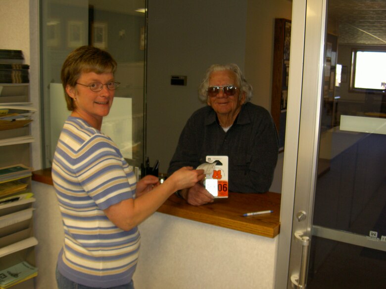 Woman in striped shirt hands man in sunglasses and long sleeve green shirt a white and orange hang tag pass through a window while both are smiling.