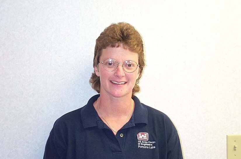A woman shoulder up is standing in front of a white wall wearing a blue polo.
