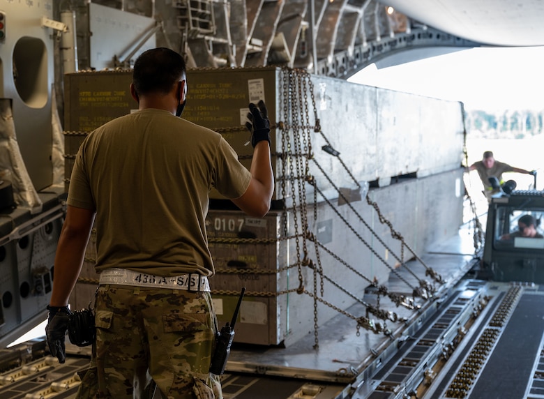 A man in uniform gives hand signals as cargo is loaded into a military aircraft.