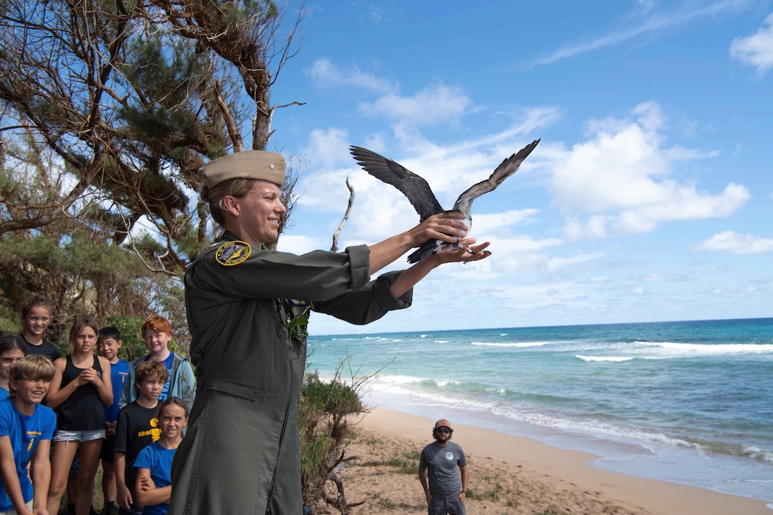 A Navy officer releases a bird while children watch on a beach.