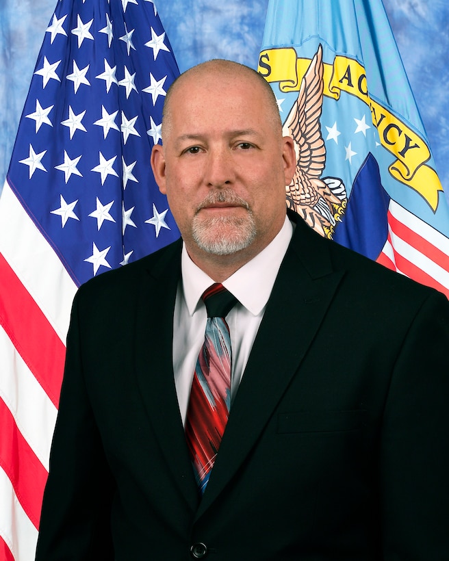 Man in black suit, white shirt and red, white, blue tie  in front of American and DLA flag for official photo