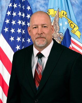 Man in black suit, white shirt and red, white, blue tie  in front of American and DLA flag for official photo