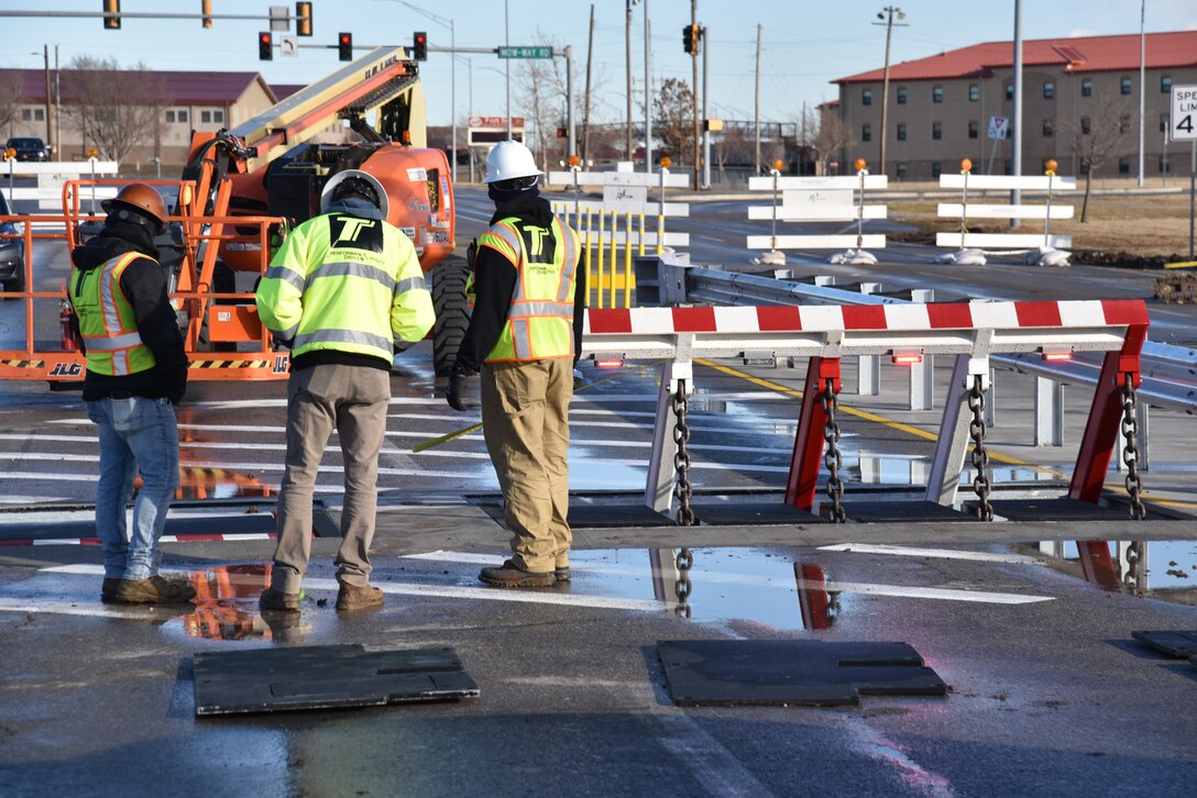 Three contractors in hard hats and safety vests stand near an activated vehicle barrier at Fort Sill , Oklahoma.