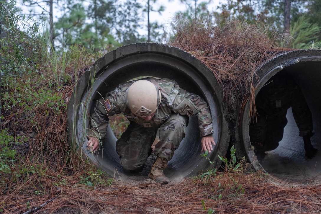 A uniformed soldier crawls through a cylinder in a field.
