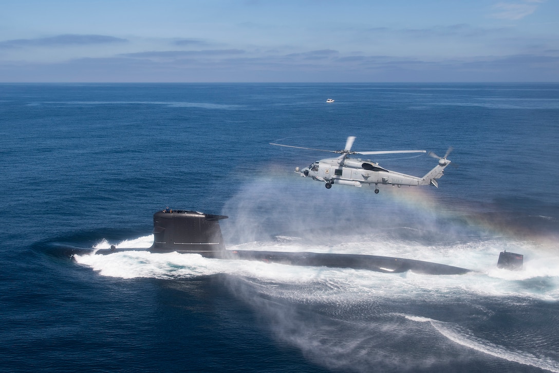 A military helicopter flies above a submarine in open water. A rainbow can be seen in the mist under the helicopter.