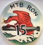 A colorized, rounded disc depicting MTBRON-15’s insignia: a red-colored dragon sitting on top of a black-colored torpedo flying in the air above ocean waves, with the words “MTB RON” and “15” on the disc.