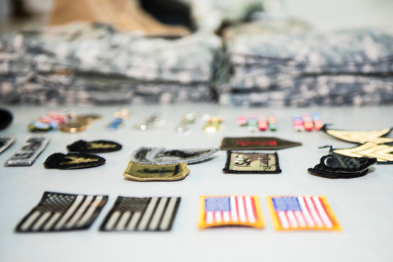 Several U.S. flag and military patches sit on a table with folded uniforms in the background.