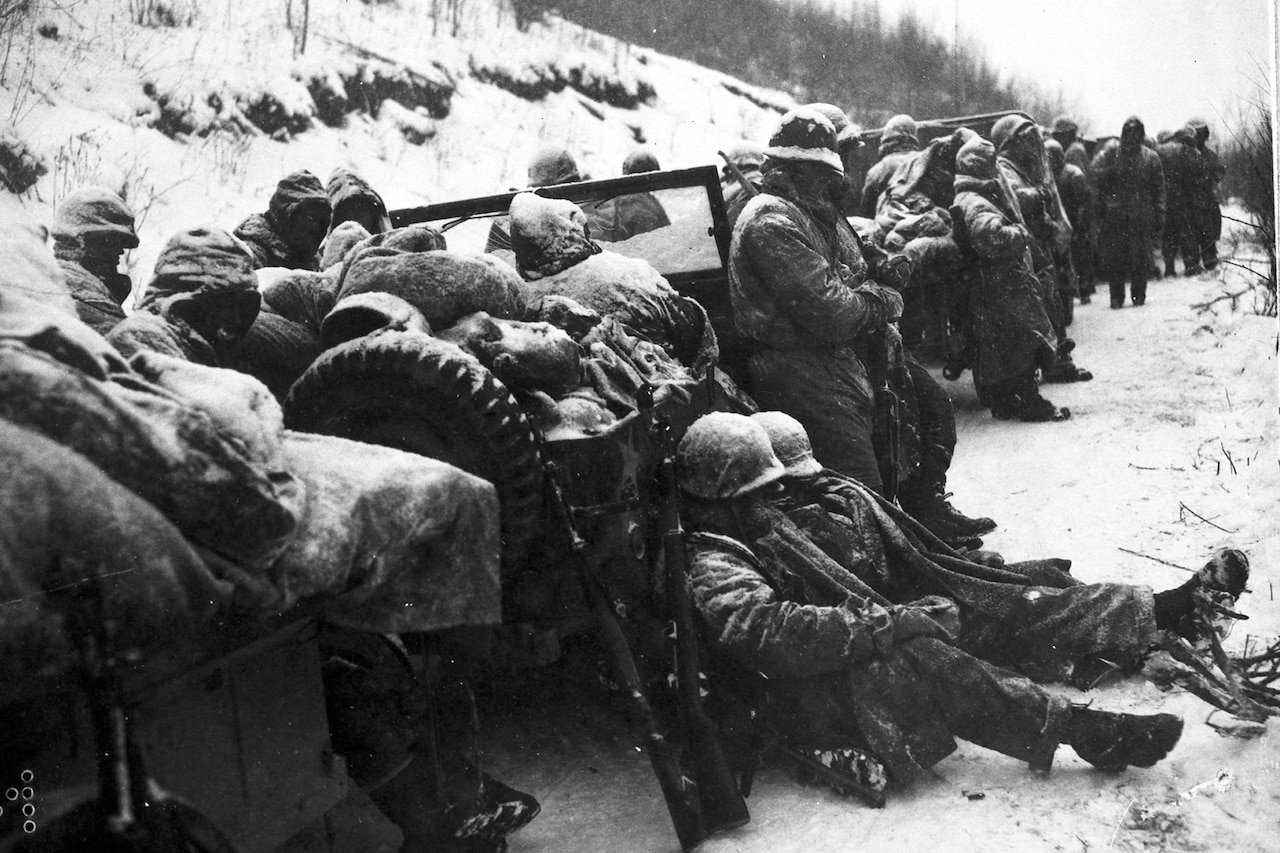 Service members in heavy winter gear relax by vehicles on a snow-covered road.