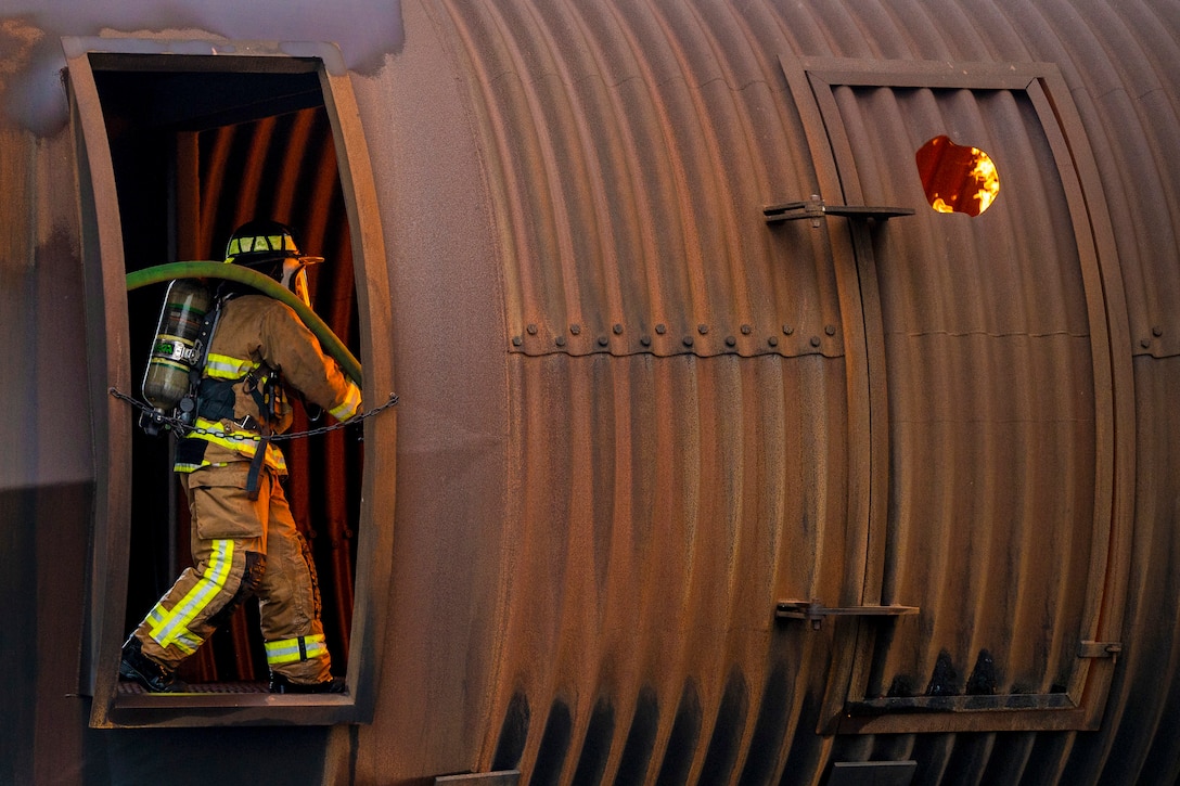 A firefighter carrying a hose moves toward a simulated fire as seen through the open doorway of a training aircraft.