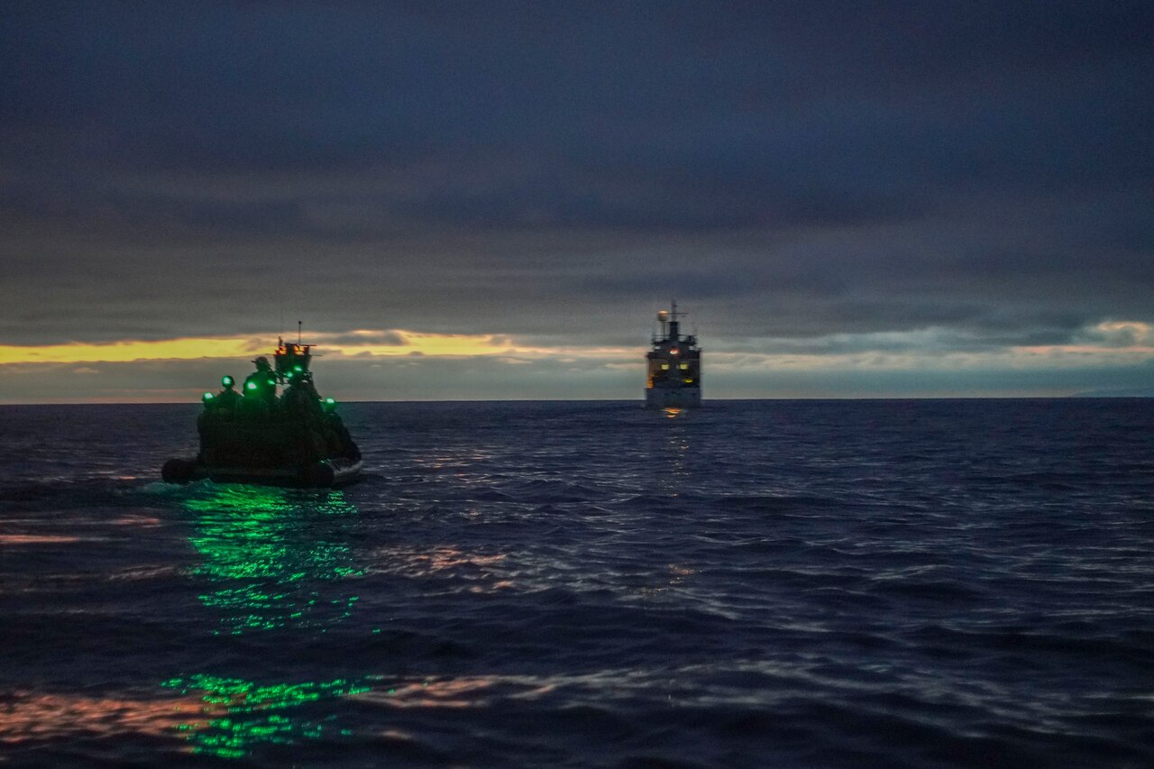 Marines in an inflatable boat approach a vessel at twilight.