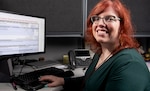 A woman sits at a desk in an office. A computer screen is in the background.