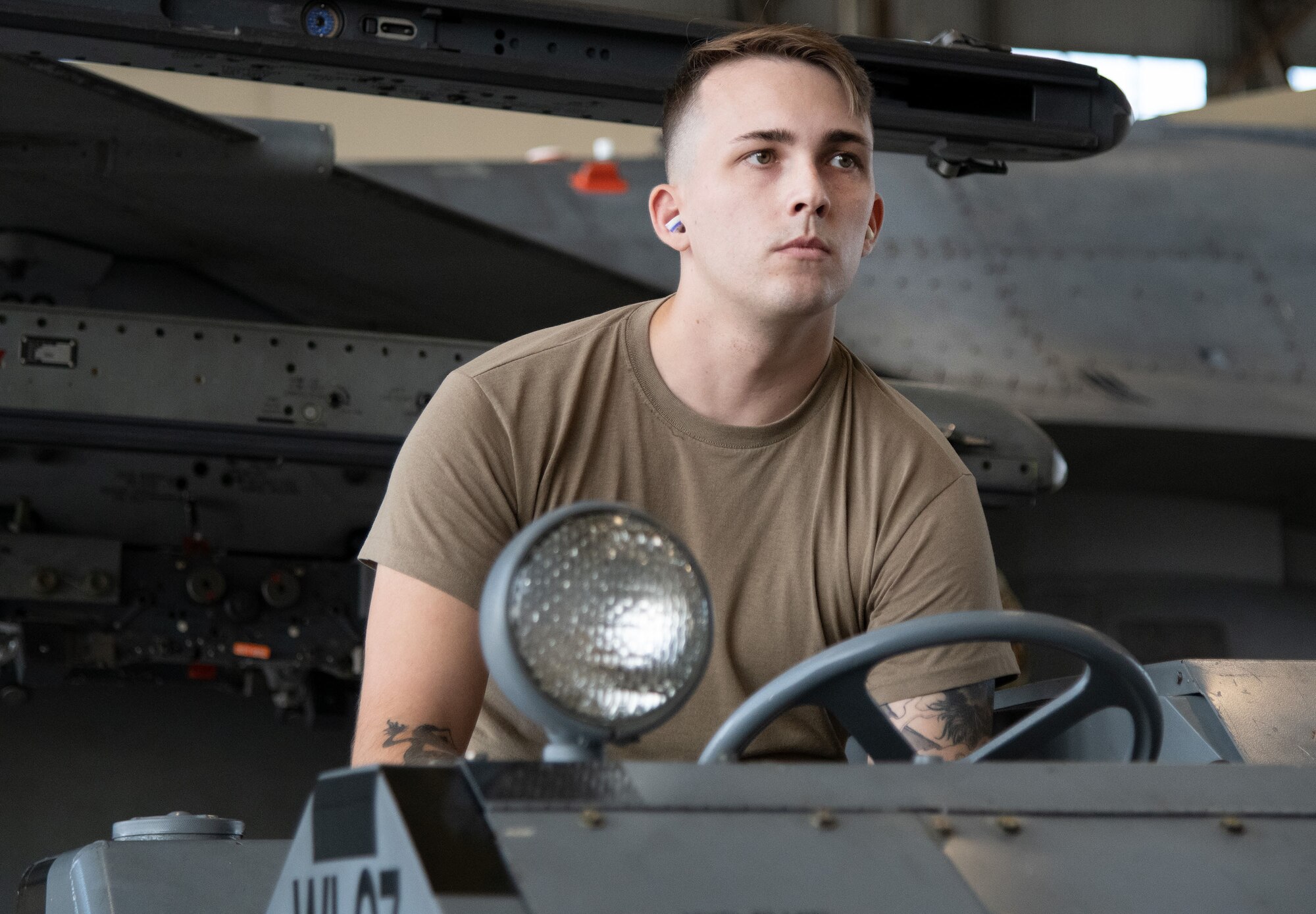 An Airman drives an AIM-120 vehicle inside a hangar, a fighter jet in the background.