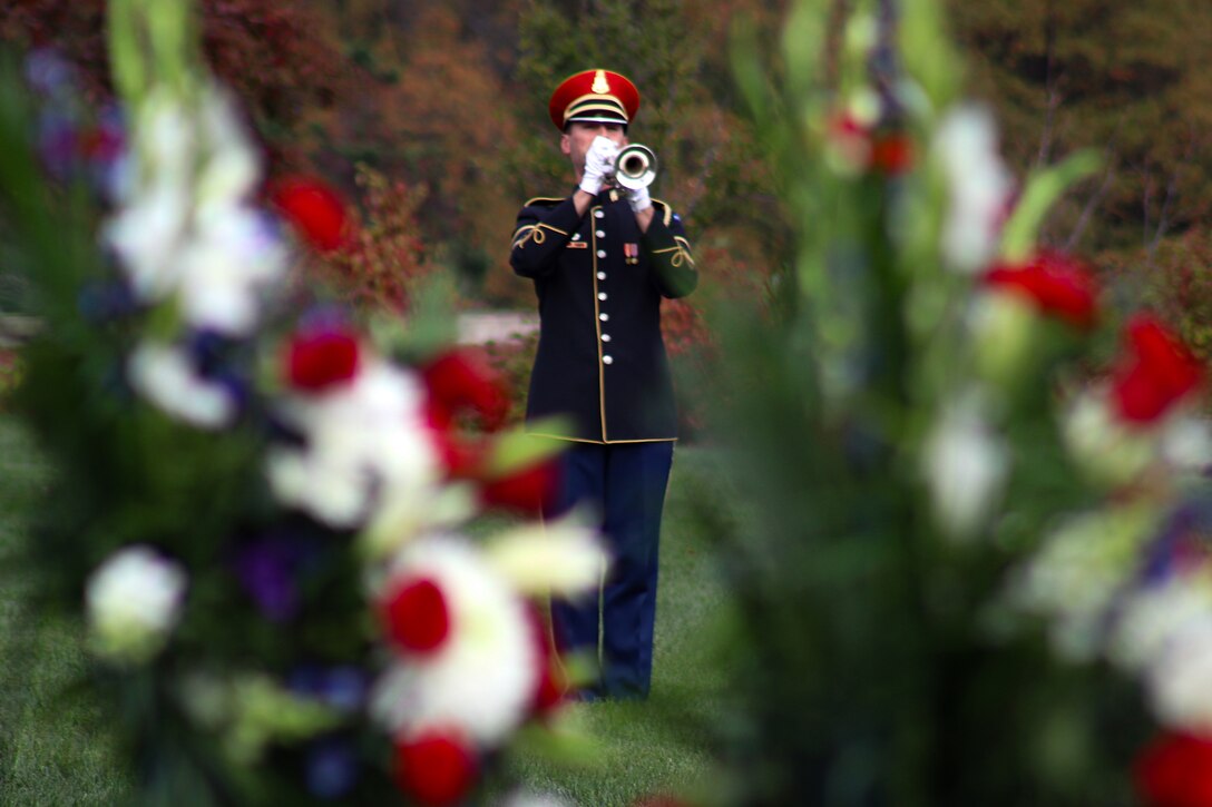 A soldier in a formal uniform plays a trumpet as seen through flowers  during a funeral.