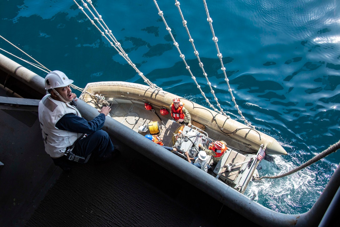 A Navy officer stands on the side of a ship above a small inflatable boat.