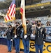 Four Soldiers in uniform stand on the field of a baseball stadium. A male and female Soldier hold flags, while the other male and female Soldiers hold guns.