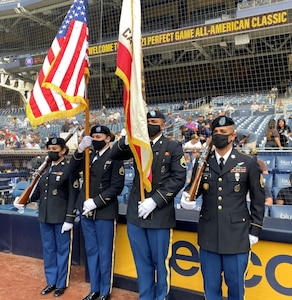 Four Soldiers in uniform stand on the field of a baseball stadium. A male and female Soldier hold flags, while the other male and female Soldiers hold guns.