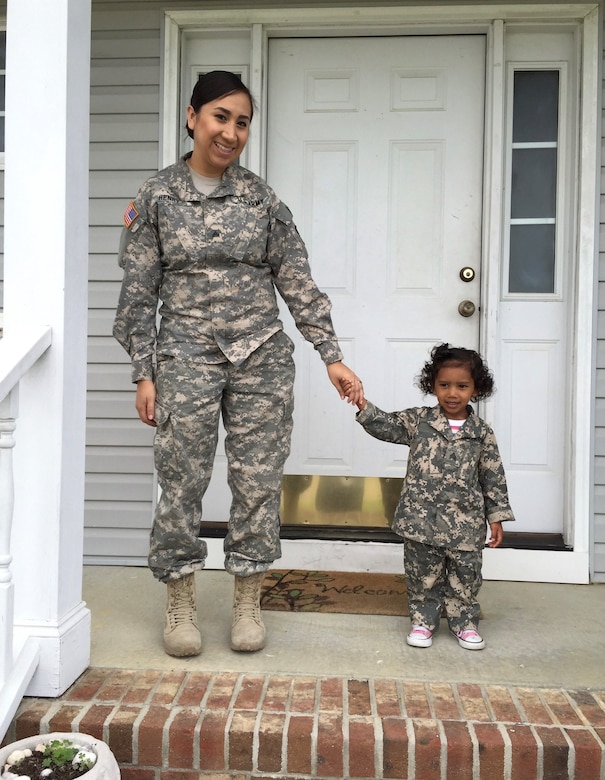 A female Soldier in uniform stands holding hands with a little girl in a Army uniform in front of a front door.