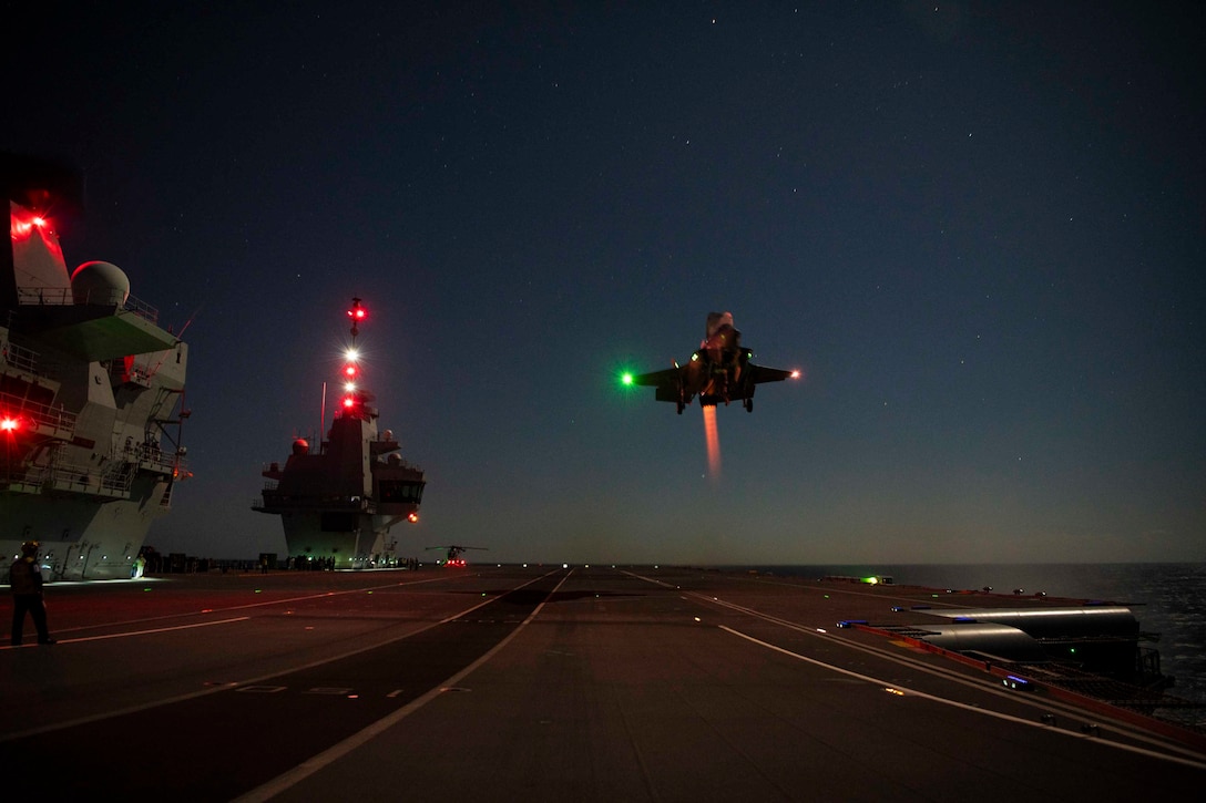 An aircraft prepares to land on the deck of a ship at night.