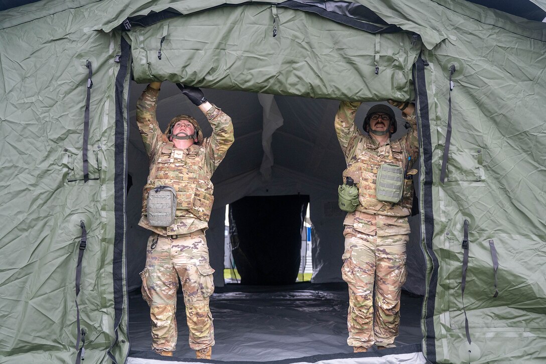 Two uniformed service members reach above them to the entryway of a tent.