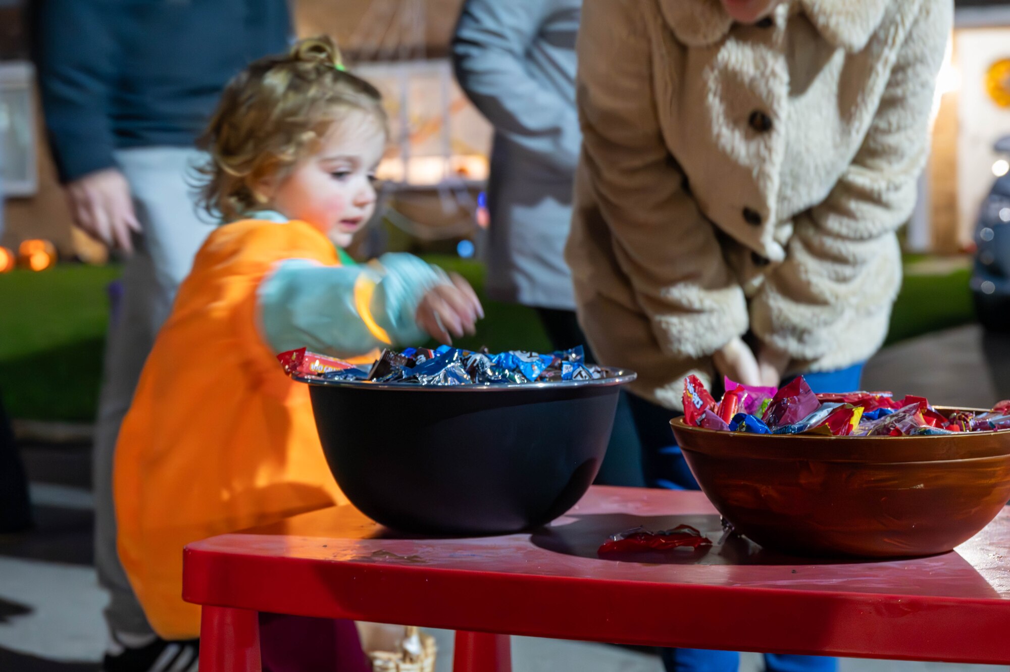 A small child dressed as a pumpkin picks up a piece of candy from a bowl