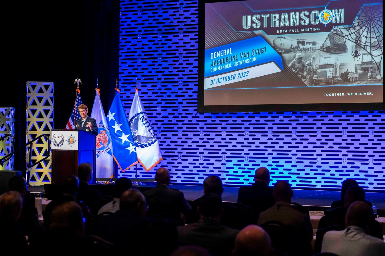 A woman in uniform speaks at a podium on stage behind a crowd sitting down.