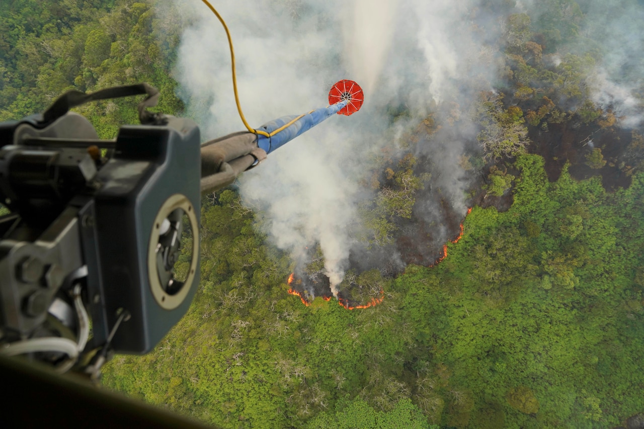 A helicopter lowers a bucket of water into a forest covered with smoke and flames.